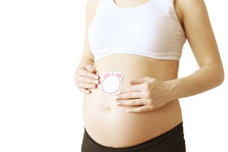 关闭孕妇穿着白色支持无缝产妇胸罩和黑色瑜伽裤与空白闹钟形状的标签在大裸露的腹部。预期概念。背景, 复制空间