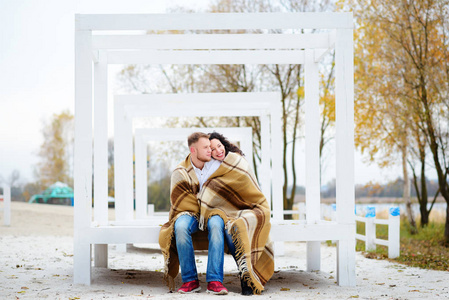 年轻夫妇拥抱和覆盖着温暖的毯子坐在秋天的海滩上