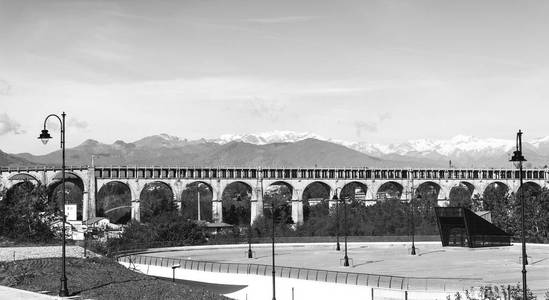 山前的汽车和铁路渡槽在积雪覆盖的阿尔卑斯背景下。在意大利山山麓的停车场。黑白图片
