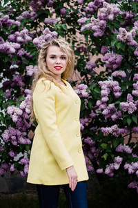 在一件黄色的大衣金发女郎站盛开紫丁香背景在春天