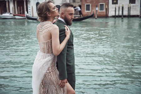 威尼斯运河附近的一对令人惊叹的新婚夫妇。优雅的女人在奢华的象牙礼服, 凌乱的发髻头发, 男人在绿色的三件西装