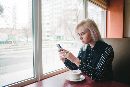 年轻的女学生坐在一个舒适的餐厅附近与一杯咖啡窗口和获得一条消息在您的手机。喝咖啡休息时间