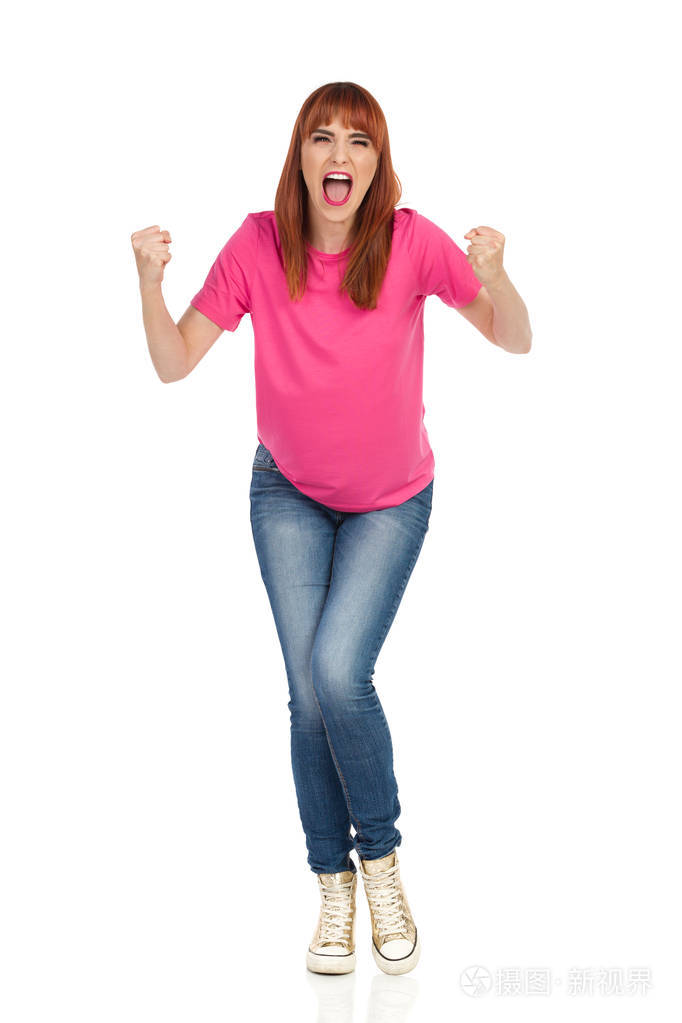 穿着粉红色 t恤衫,牛仔裤和金色运动鞋的愤怒的年轻女子紧握拳头大喊.