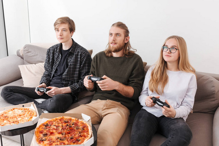 一群朋友坐在沙发上, 在家里玩电子游戏。两个男孩和女孩的肖像吃比萨饼和玩游戏隔离