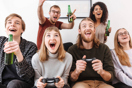 关闭照片的兴奋的朋友坐在沙发上和花时间在一起玩电子游戏和喝啤酒在家里