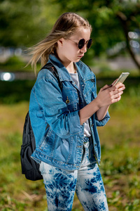 一个十几岁的女孩手持智能手机。夏天在自然放学后。穿着牛仔服在背包后面。将邮件写入家长