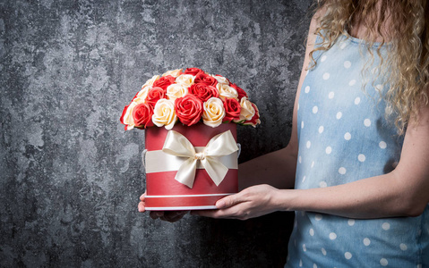一个穿着蓝色连衣裙的女孩在帽子盒里捧着一束红白相间的玫瑰花。深灰色背景。特写