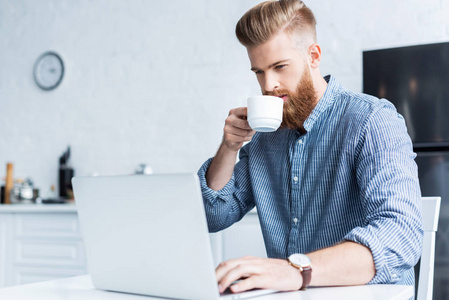 英俊的胡子年轻人喝咖啡和使用膝上型电脑在家中