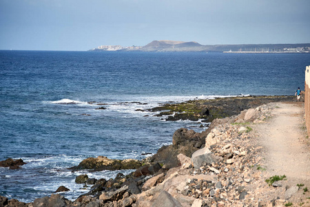 豪华海滨。海的岩石岸边, 海浪敲打岩石。一个带着蓝色背包的游客在后台漫步。与口岸的城市在距离是可看见的