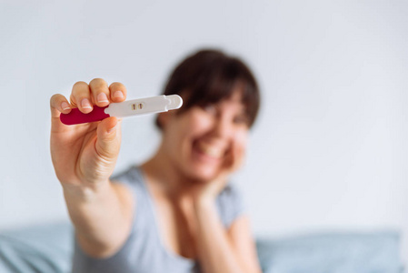 年轻快乐的女人坐在床上看阳性妊娠测试。背景模糊