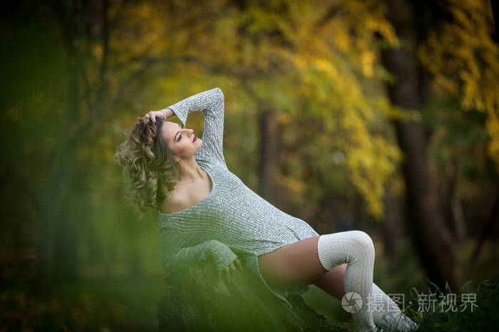 长腿坐在一个树桩在一个秋天的场景与感性的女孩。长腿有吸引力的金发卷发轻松在秋天的公园。时尚的年轻女子，在森林里一个树桩上摆姿势