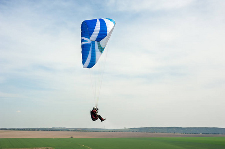 在晴朗的日子里, 蓝天上的滑翔伞。春季在田野和河谷下的蓝色滑翔伞飞行