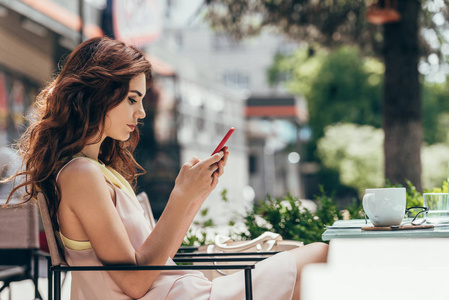 年轻女子使用智能手机的侧面视图坐在桌边咖啡馆里喝咖啡