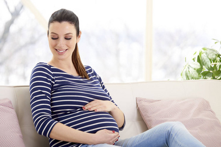 美丽的怀孕妇女与手在她的婴儿颠簸坐在沙发上, 在家放松