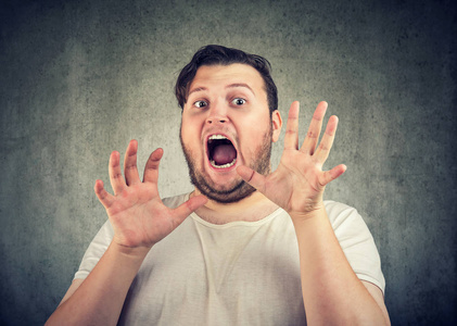 胖胖的休闲男子手放在脸上, 惊恐地尖叫着看着在灰色背景下的相机照片