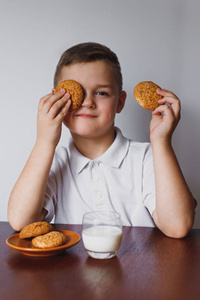 一个小男孩拿着燕麦饼干。玩饼干的乐趣。桌子上有美味的饼干和新鲜的牛奶。一个小学生对着白色墙的肖像