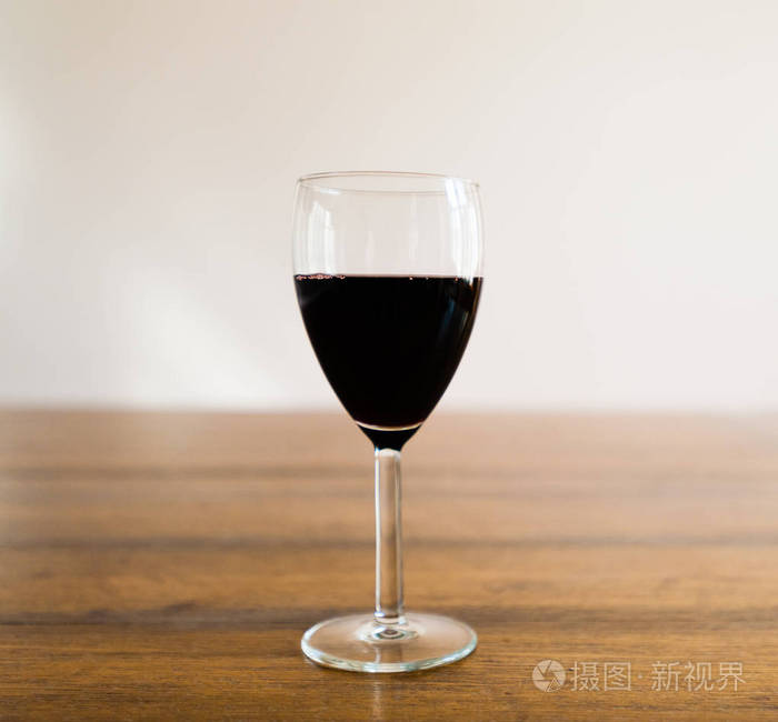 一杯半满的黑红葡萄酒站在木桌上, 对着白色的墙在模糊的背景下