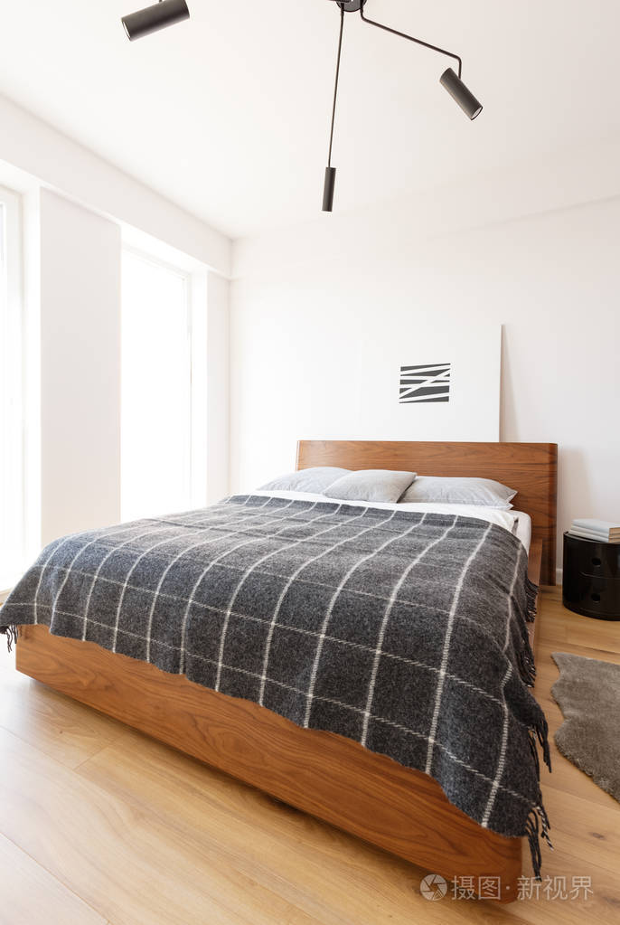 图案深色毛毯在木床与海报在白色简单的酒店卧室内部.真实照片