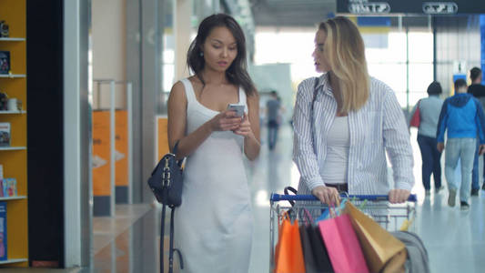 两个年轻女人用智能手机和交谈购物车