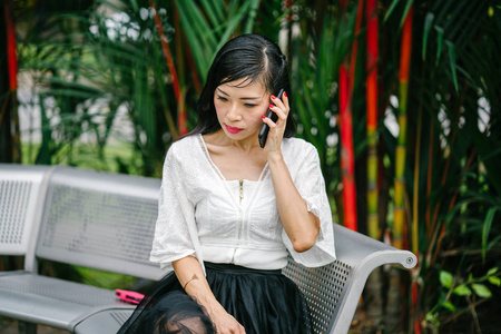 一位亚洲妇女坐在一个金属长凳上, 上面有一台带有智能手机的背景树。她是中年, 瘦, 穿着办公室服装