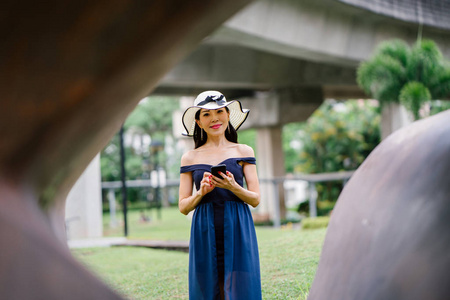 一个时尚的亚洲妇女的肖像在蓝色的礼服和白色的帽子公园与智能手机