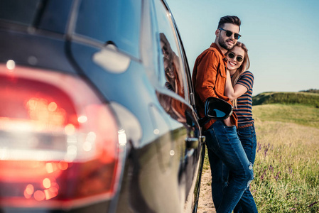 快乐的时尚情侣在太阳镜下站在乡村草地附近的汽车