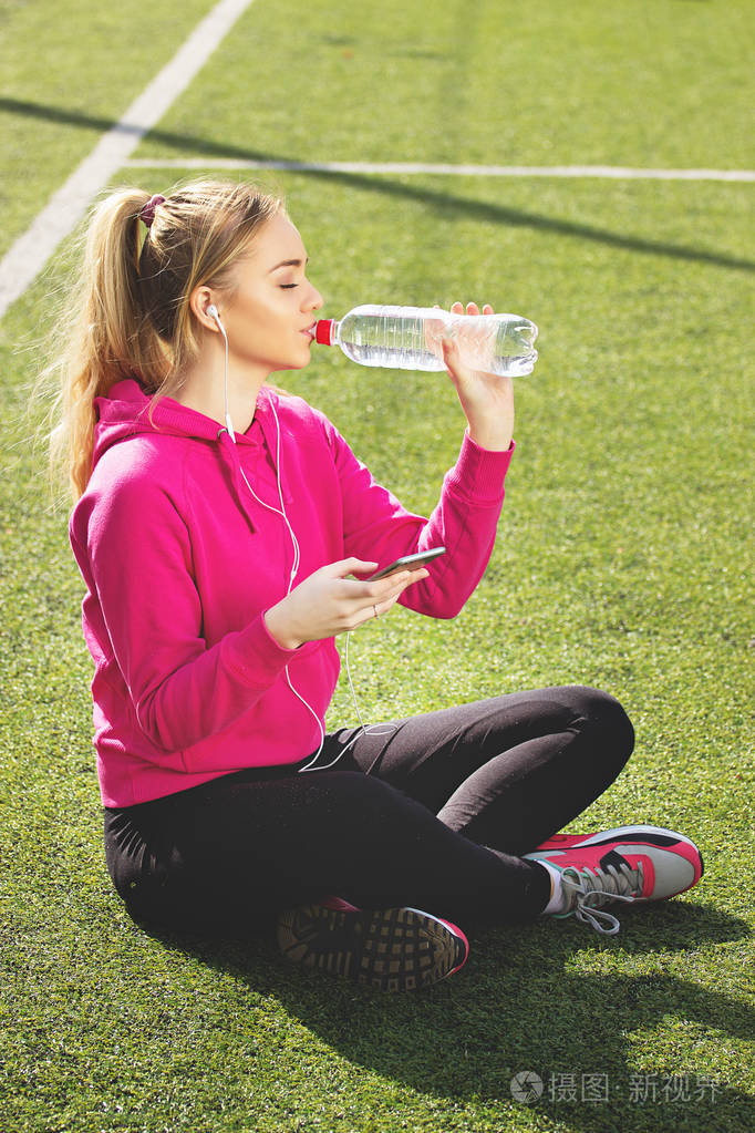 渴了年轻漂亮的金发健身女孩喝水在体育场上夏季体育活动绿色球场草地