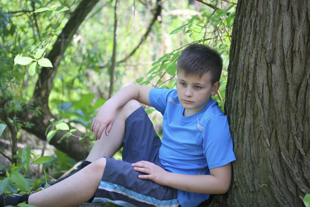 一个十几岁的年轻人在公园散步时为摄影师拍照。坐着, 倚在树上