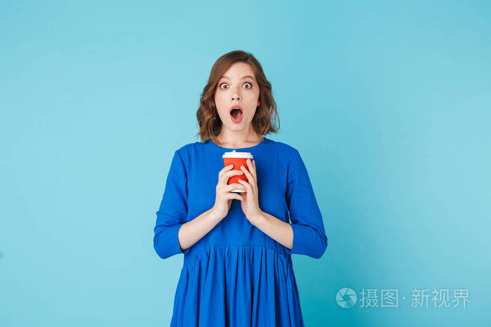 蓝色礼服的年轻女士的照片站在杯咖啡去和惊异看着在蓝色背景上的相机