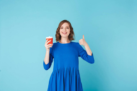 年轻女士的肖像站在一杯咖啡, 在蓝色的背景下去。漂亮的女孩高兴地显示大拇指向上的手势, 而看着相机
