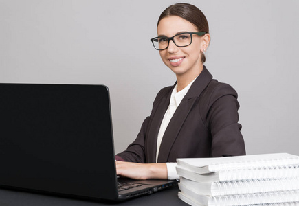 开朗的商业妇女坐在办公桌上, 文件和书籍, 并使用笔记本电脑
