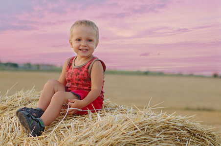 小男孩的肖像坐在一个麦田里的干草堆上的一个夏天的帽子。美丽的日落