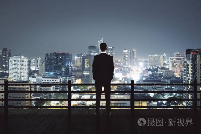 在屋顶的年轻商人看着照亮的夜晚城市的背面视图.研究与成功理念