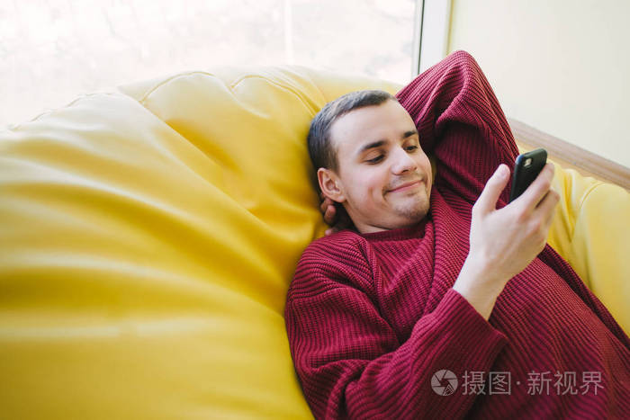 积极年轻的男同学躺着,看着他的智能手机.后在舒适的椅子上的学习.