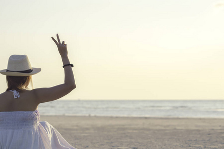 在日落时, 坐在热带海和海滩上的快乐女人的背面显示两个手指或胜利标志。年轻女子享受生活假期和放松在天堂岛的复古色调