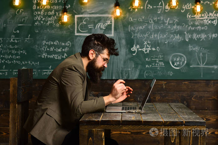 有胡子的人在教室里工作笔记本电脑。长胡子的人用电脑在黑板上。西装商人在校服务台使用笔记本电脑。戴眼镜的科学家在天才的脸上。新技术