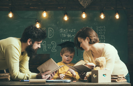 在家上学的概念。父亲和母亲看书, 教他们的儿子, 黑板上的背景。家庭关心他们的儿子的教育。孩子在研究生盖帽喜欢听故事