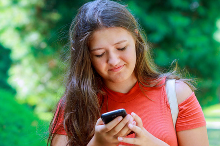 一个美丽的少女的肖像在一个绿色不集中背景的公园里, 在智能手机上键入信息