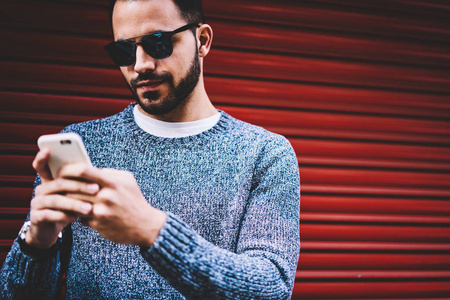 英俊的年轻人发送短信聊天的智能手机站在城市设置背景, 时髦的家伙检查通知手机更新应用程序花费空闲时间户外