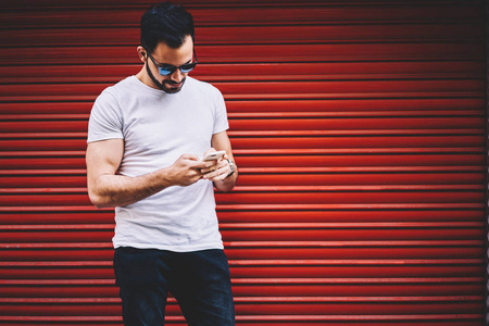 英俊的时髦家伙银行业务通过应用手机站在户外的宣传背景下, 年轻时髦的着装男子智能手机在社交网络中与追随者聊天的短信