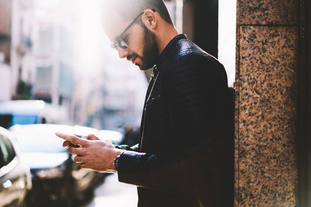 沉思的时髦的家伙阅读收入短信和发送反馈通过智能手机站在户外, 年轻男性观看视频在线电话连接到4g 互联网网络在城市街道上