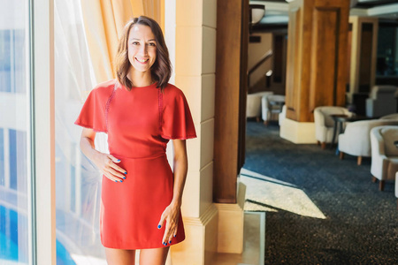 一个穿着红色连衣裙的年轻女孩站在窗边。在酒店的大窗户上的一个女人的肖像。酒店大堂的女孩
