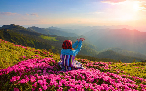 女孩坐在草坪上, 上面覆盖着粉红色的花朵, 在高山上看着天空, 夏日里有云彩和太阳。对自然爱好者的好看法