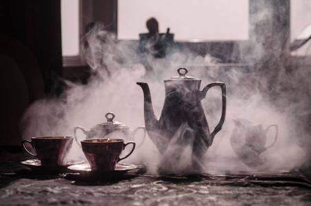 食品背景茶和咖啡的主题。老的古董陶瓷茶或咖啡壶杯壶和糖杯带灯和烟雾的黑暗背景上。轮廓的陶瓷茶壶和杯