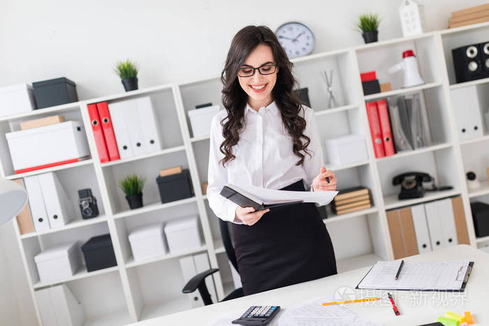 一个漂亮的年轻女孩站在办公桌旁, 手里拿着一支钢笔和一本笔记本.