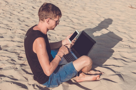 与便携式计算机和移动电话在海滩上的年轻男子。做