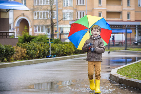 在多雨的夏季公园玩耍的小男孩。儿童与彩虹伞，防水大衣和靴子在水坑里跳和泥在雨中。孩子走在秋日的阵雨户外乐趣的任何天气