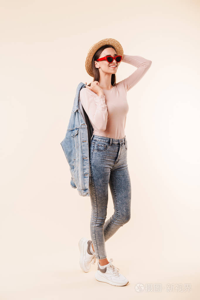 全长肖像的一个快乐的年轻的黑发女孩在草帽和牛仔夹克姿势, 而站着