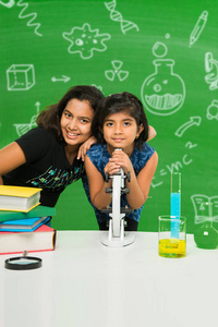 2 小印度女孩学习科学或生物显微镜在课堂上使用在有科学涂鸦用粉笔绘制的绿色黑板背景