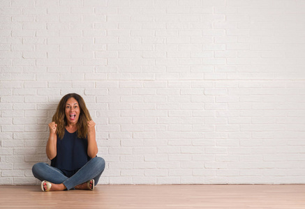 中年拉美裔妇女坐在地板上的白色砖墙庆祝惊讶和惊讶的成功用双臂举起和睁开眼睛。赢家理念